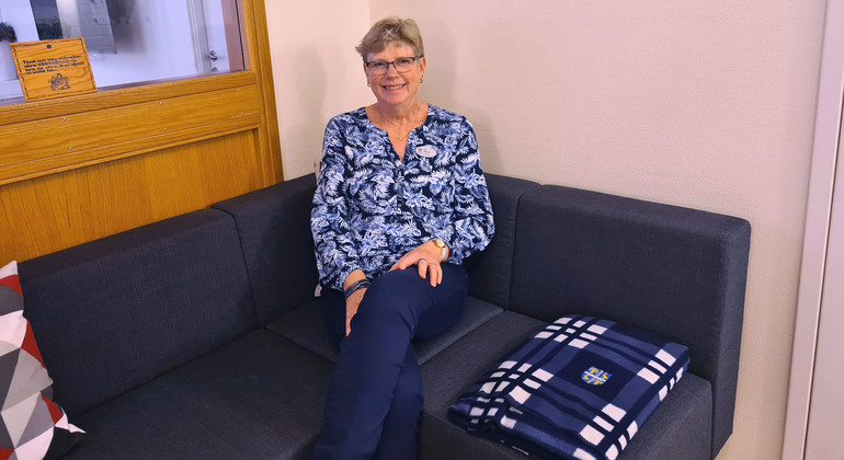 Eva Olsson sitter på en blå soffa. Hon har benen i kors och ler mot kameran. Bredvid sig har hon en blåvit filt. 