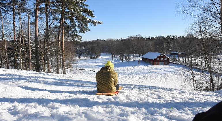 Fotograf: herrljunga Kommun. Foto visar person i grön jacka samt gul mössa i Harabergsbacken. Snö på marken och utsikt över stoltamaderna samt vagnsmuseet. Väder: Solig dag med blå himmel.